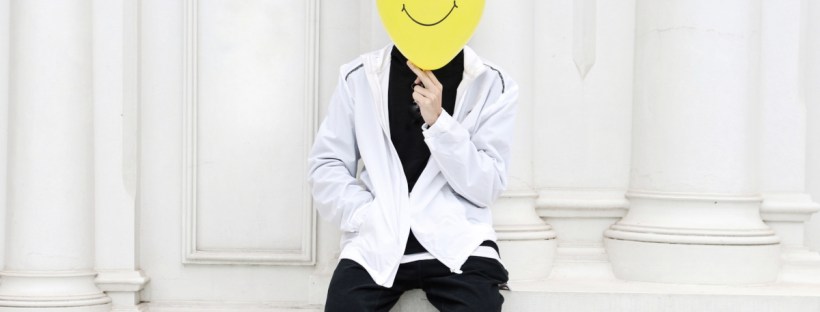 A person in a black and white outfit holds a balloon over their face. A smiley face is drawn on the balloon.