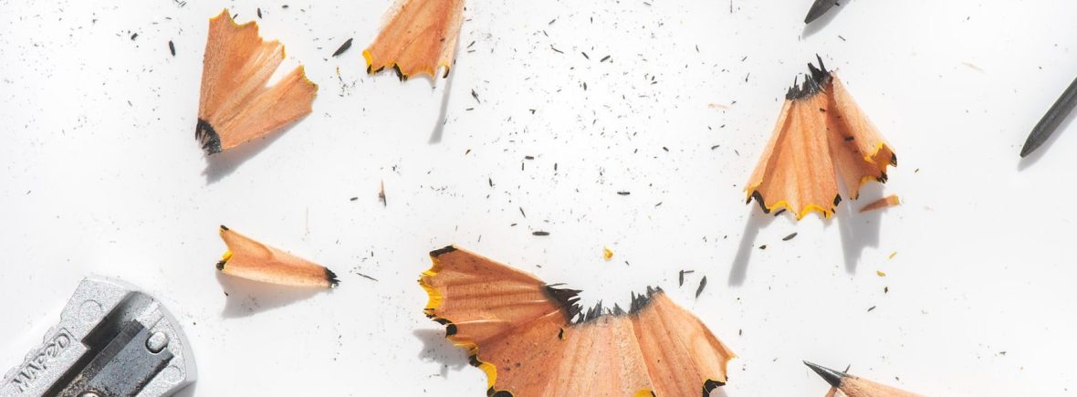 Pencil shavings litter a white background. A sharpened pencil, three slivers of led, and a pencil sharpener are also in the frame.